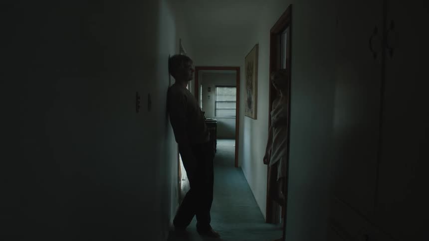 A man leans against the wall looking at a woman in the corridor