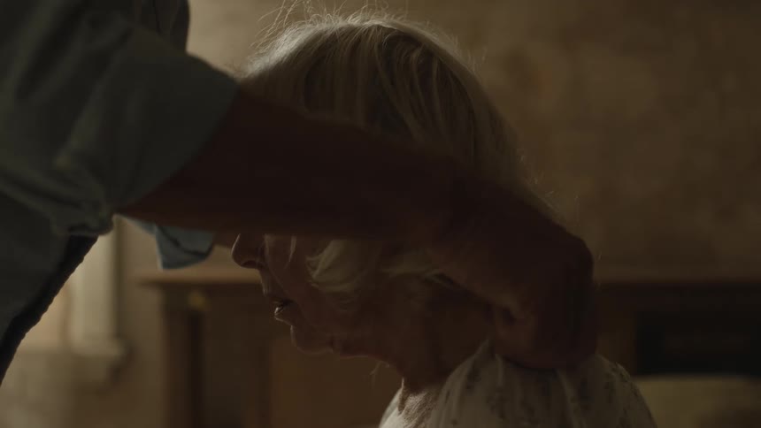 An elderly man helps an elderly woman get dressed while she sits on the bed