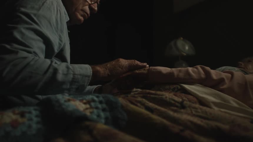 Elderly person sitting on the bed massaging the hands of his wife who is lying down