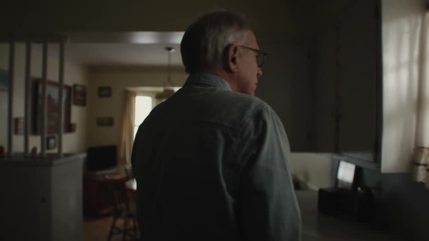 Man with glasses and light blue shirt walks in the kitchen to take a glass of water