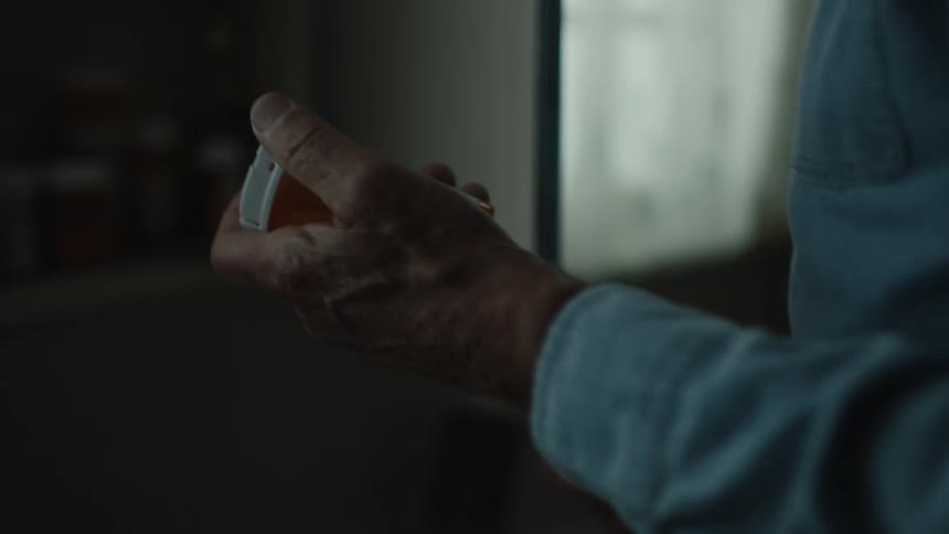 An elderly man with glasses wearing a blue shirt takes medicine in the bathroom