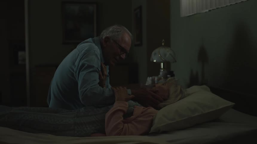 An elderly man sits on the bed where his wife is lying he caresses her soothingly as she reaches up to him as they gaze at each other