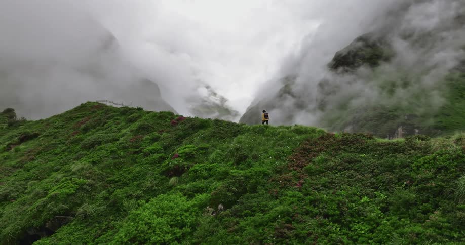 A man hiking on top of a mountain path covered in grasses as there are fog covering the atmosphere