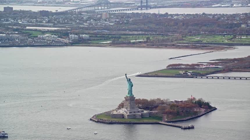 The statue of liberty holds her torch high as she overlooks new york harbor as traffic moves on the streets
