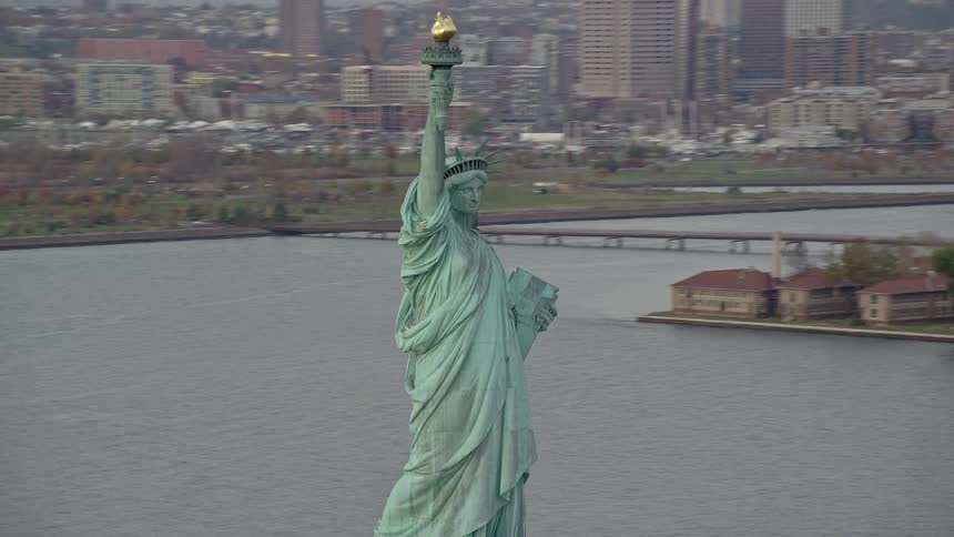 The statue of liberty holds her torch high as she overlooks new york harbor with a view of towers and skyscrapers along the hudson river as boats sail on the water