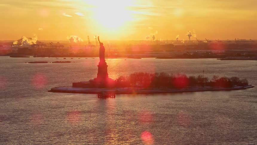 The statue of liberty stands tall on liberty island overlooking the hudson river as the sun shines brightly on the horizon
