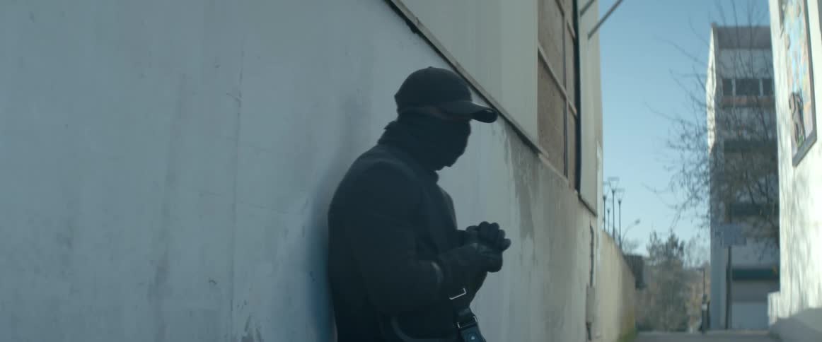 A man standing beside the street carrying a cross bag wearing a mask a baseball cap and all black outfit