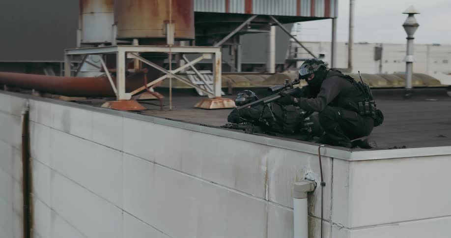 Swat team sniper crouches next to spotter on rooftop and takes aim