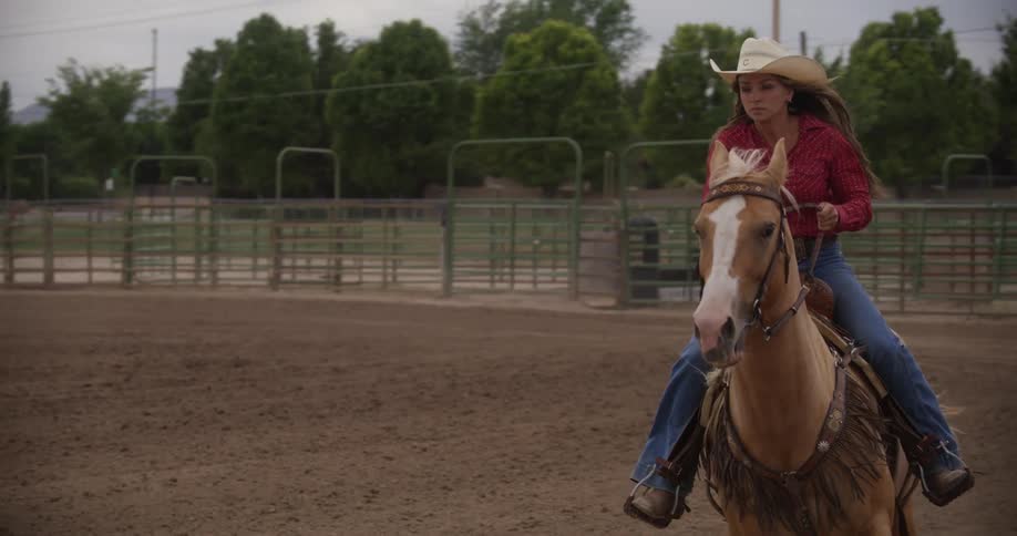 A blonde woman in a hat is riding a horse on a ranch with fences