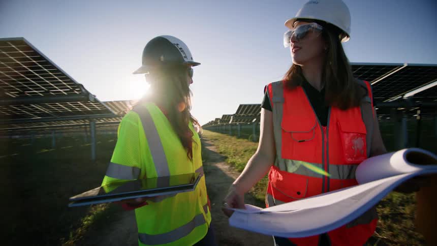 Two women in a solar field wearing safety hats and vests while inspecting the panels