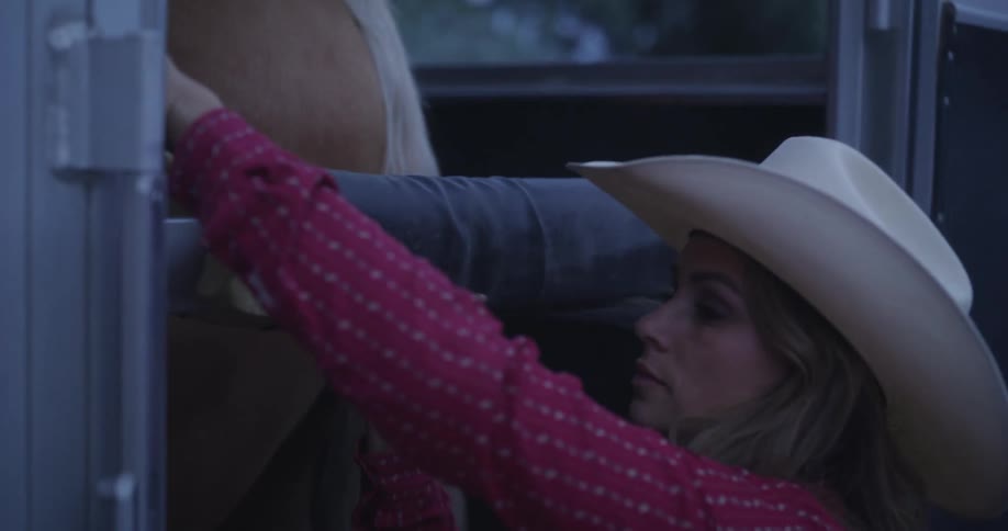 A woman wearing a cowboy hat secures a horse in a trailer