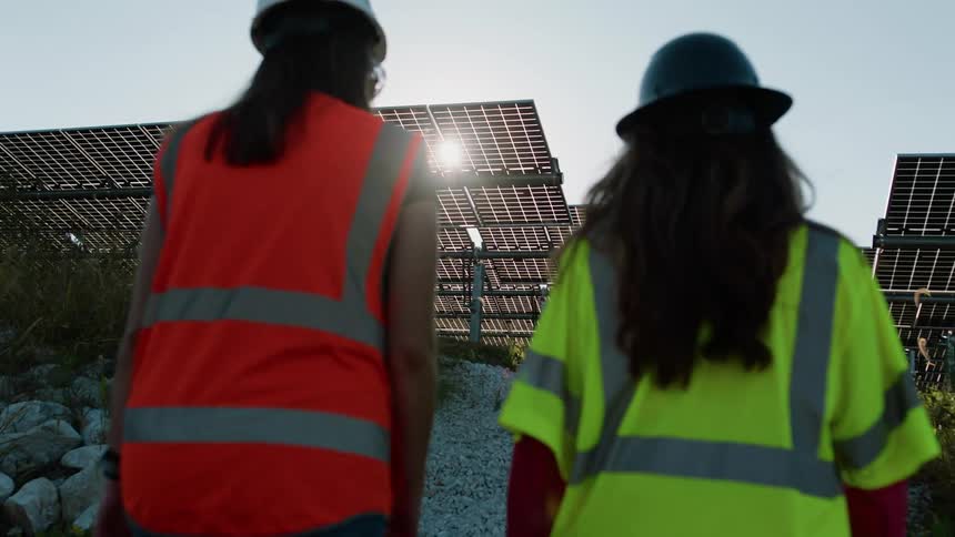 Two female construction workers inspecting solar cells on a solar farm wearing hard hats and neon vests