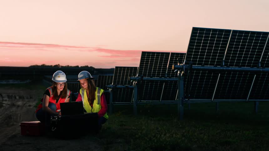Two female utility workers are sitting with a computer near solar panels at sunset