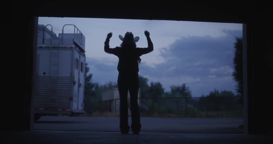 A silhouette of a woman opening a garage door and a cabalo is eating next to a fence