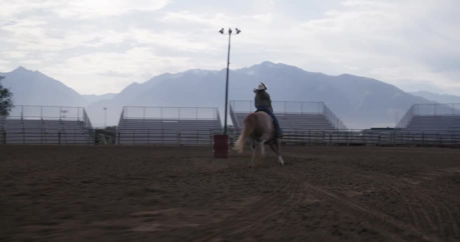 A horse races around a barrel in a rodeo arena pen in front of empty bleachers with a female rider on its back under a cloudy white sky at dusk