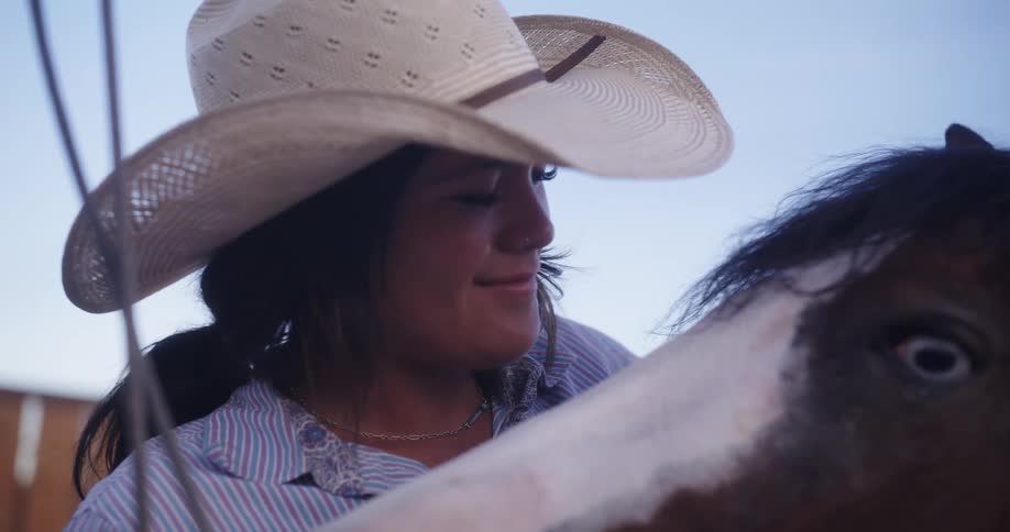 A woman in a white cowboy hat smiles as she slowly pets a horse as the horse moves its head close to hers on a clear day