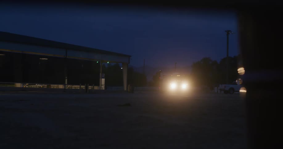 A heavy duty pickup truck hauling a horse trailer turns into a dirt lot and drives along the side of a large covered riding arena with its headlights illuminating the dark ranch at night