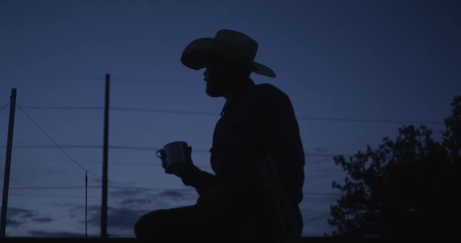 Cowboy sitting and drinking from his mug under a dark dawn sky