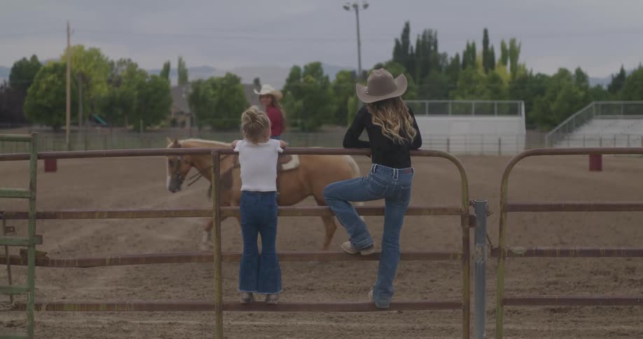 Two girls stand on a fence and watch a woman ride a horse in an arena