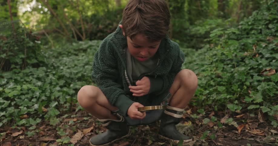Boy crouched on the ground in a forest holding a magnifying glass and observing