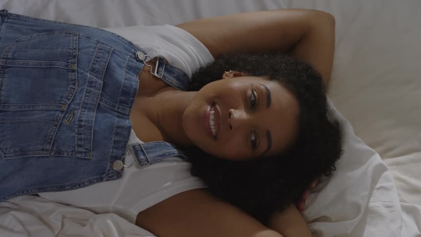 A woman wearing overalls is lying on a bed and smiling and talking to someone