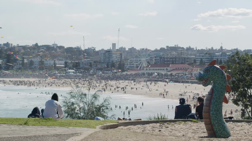 A hill with a dragon statue and some people and below there is a beach with several people bathing in the sea