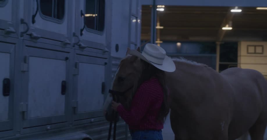 A cowgirl leads a horse around from the back to the side of a horse trailer and ties it offin a dimly lit area of an indoor riding arena at night