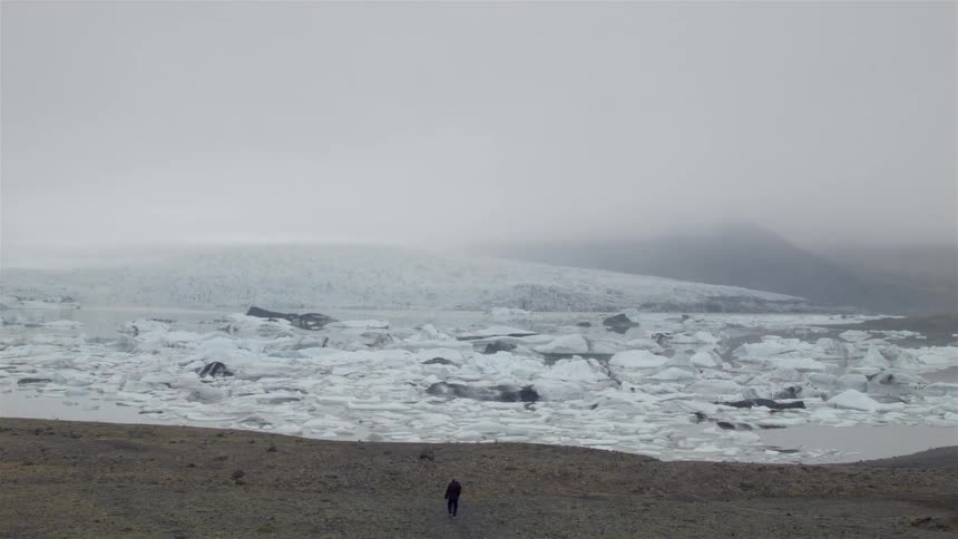 Two people walk on sand towards an iced over body of water