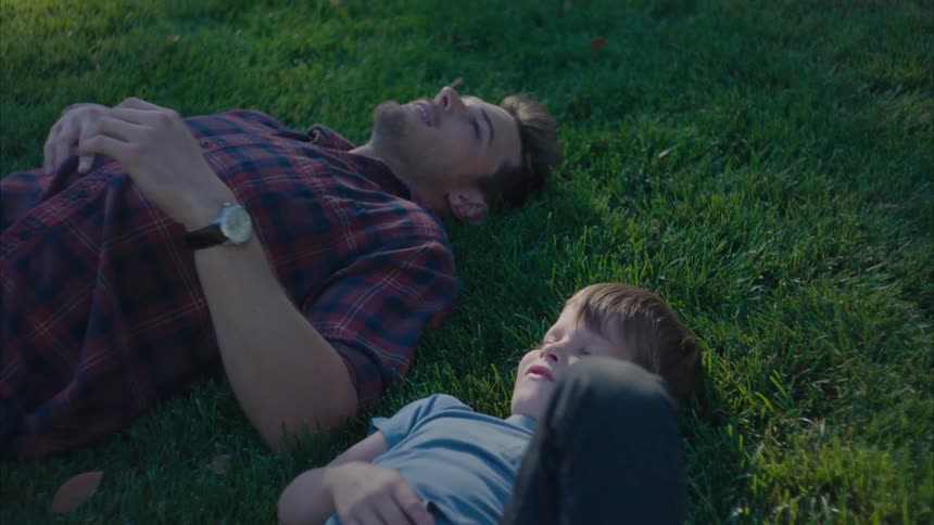 Father and son fist bump while lying on their backs on the grass