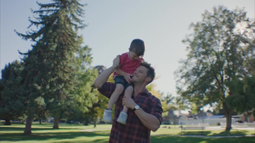 A boy smiling on the shoulder of his father in a park during the day