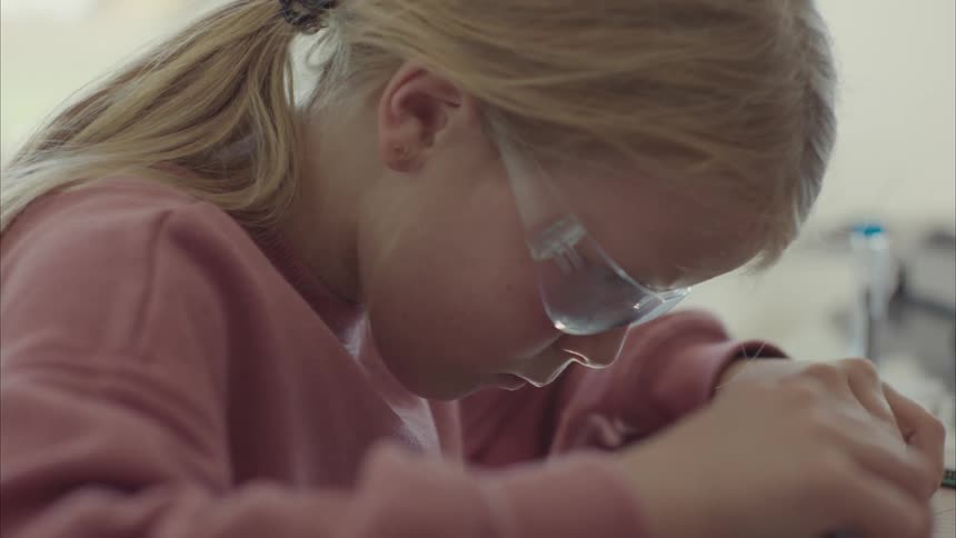 A little blonde girl with glasses is repairing a circuit board with a tool
