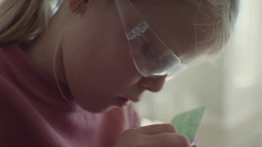 Blonde girl wearing safety glasses examines small circuit board