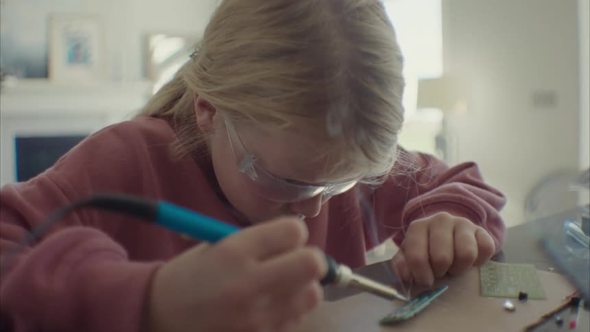 A little blonde girl welding something and is wearing glasses and a soldering iron