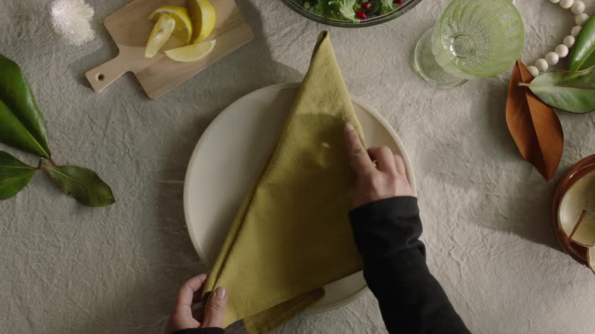 A person setting up a dining table by placing a napkin on a plate