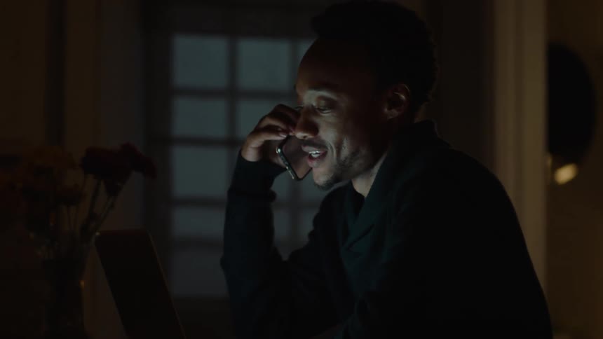Man on a computer smiles after taking a mobile call indoor at night