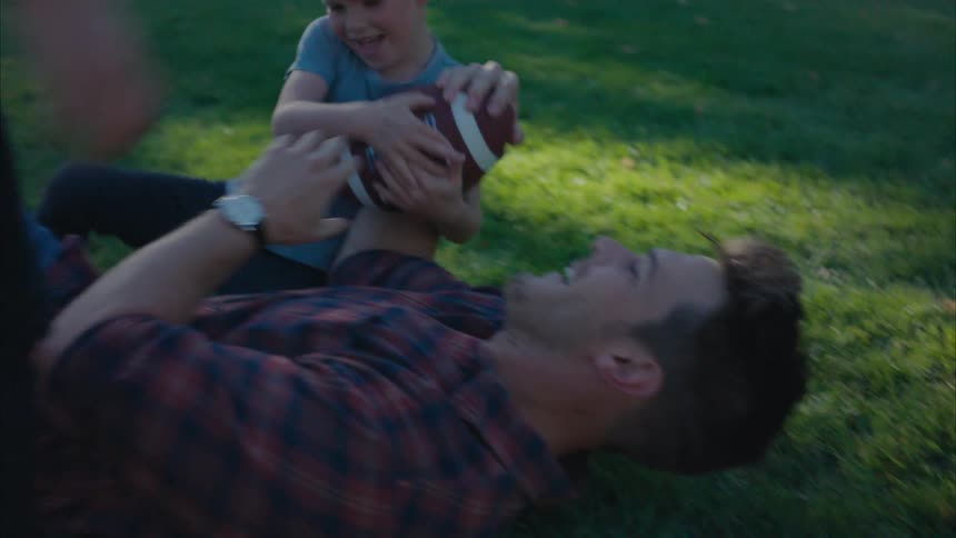 Father playing with his children falling to the ground as he tries to grab the american football from them