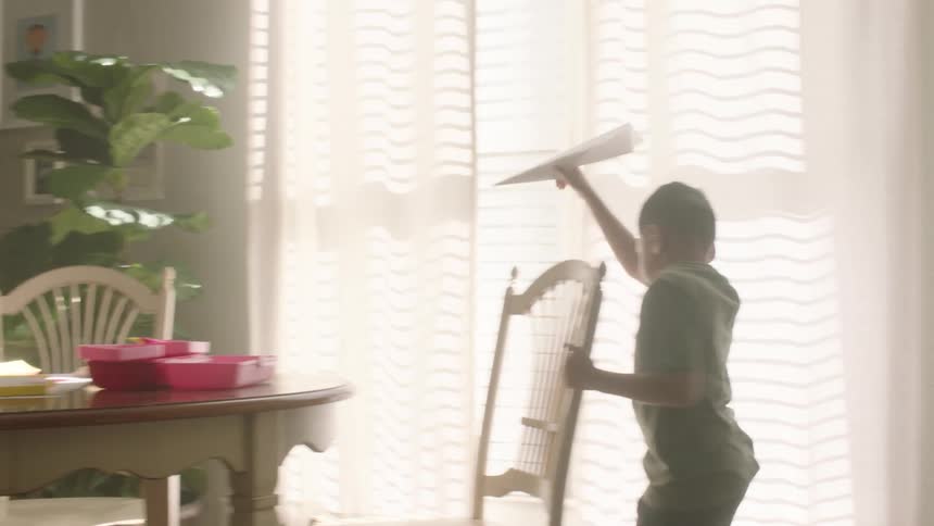 A little boy running with a paper airplane in his hand and an adult is at a table gluing something together