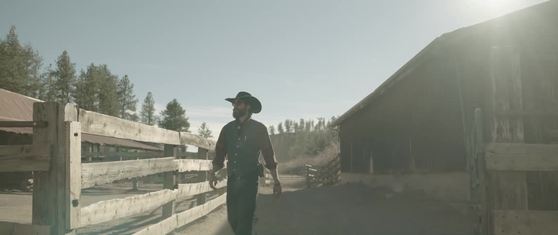 Cowboy walks between fence and barn on sunny day