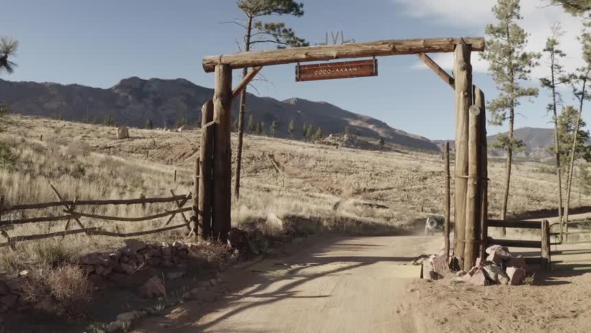 A jeep drives on the dirt path at the entrance to a dude ranch picking up speed with a mountain view in the distance as the sun beams down