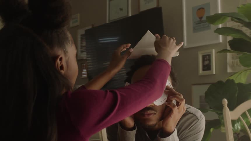A man puts a paper mask on his head and a girl puts a paper boat on the mans head
