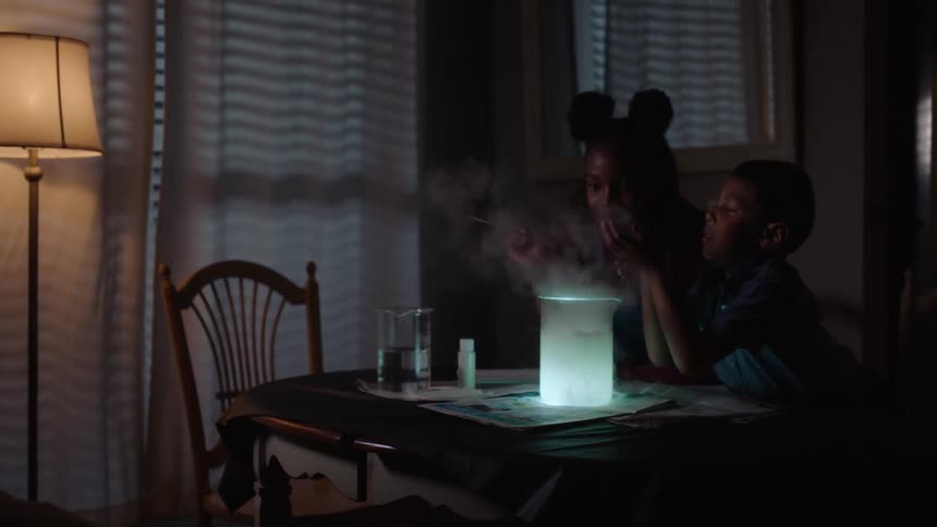 A girl wearing mickey mouse ears and a younger boy do science experiments on a dinner table