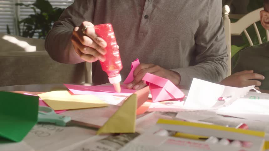 A man sitting is applying glue to a piece of paper and next to him is a little boy