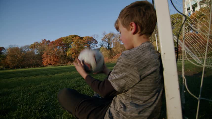 Boy sitting against a goalpost on a grassy field tossing a ball up with his hands