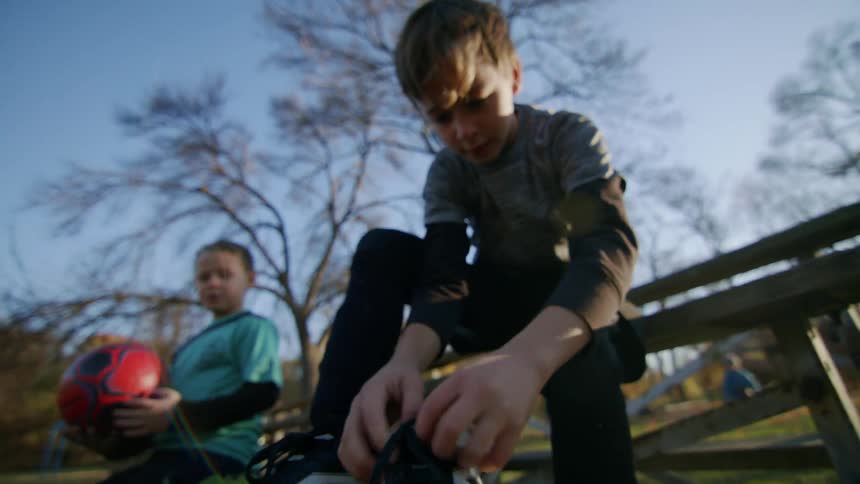 A boy tying his boot laces sitting on a wooden bench