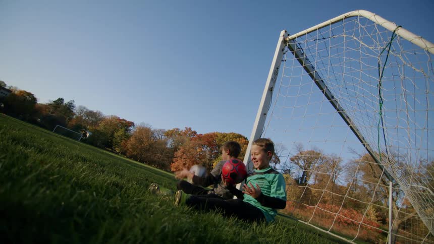 Two boys sits on ground near soccer goal and play with soccer balls