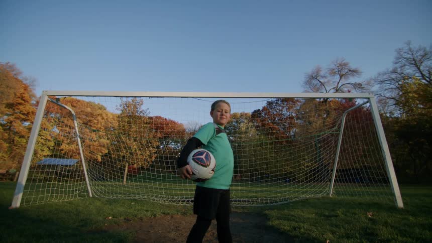 A boy confidently posing for the camera in front of the soccer goal holding a ball