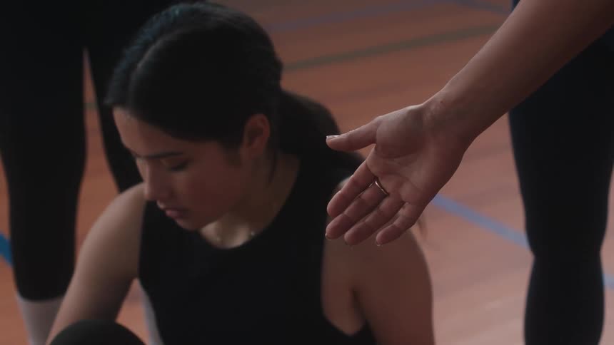 Teenage girl sits on gymnasium floor and reluctantly accepts a hand up