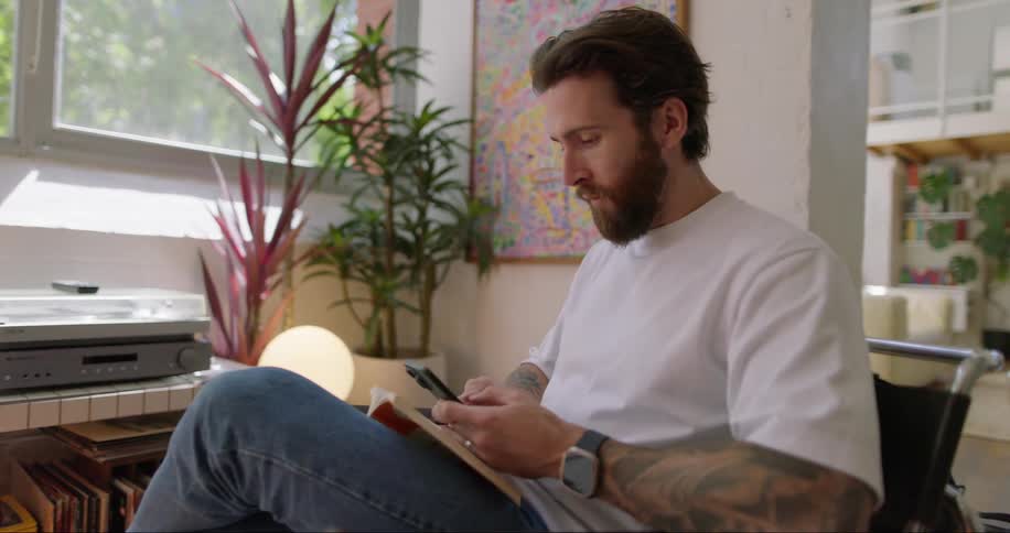 A man sitting reading a book and using his cell phone