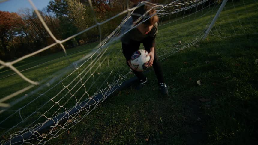 A boy picks up a soccer ball from the net at sunset on a soccer field