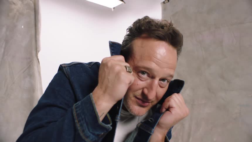 A man holding the collar of his denim jacket in a studio